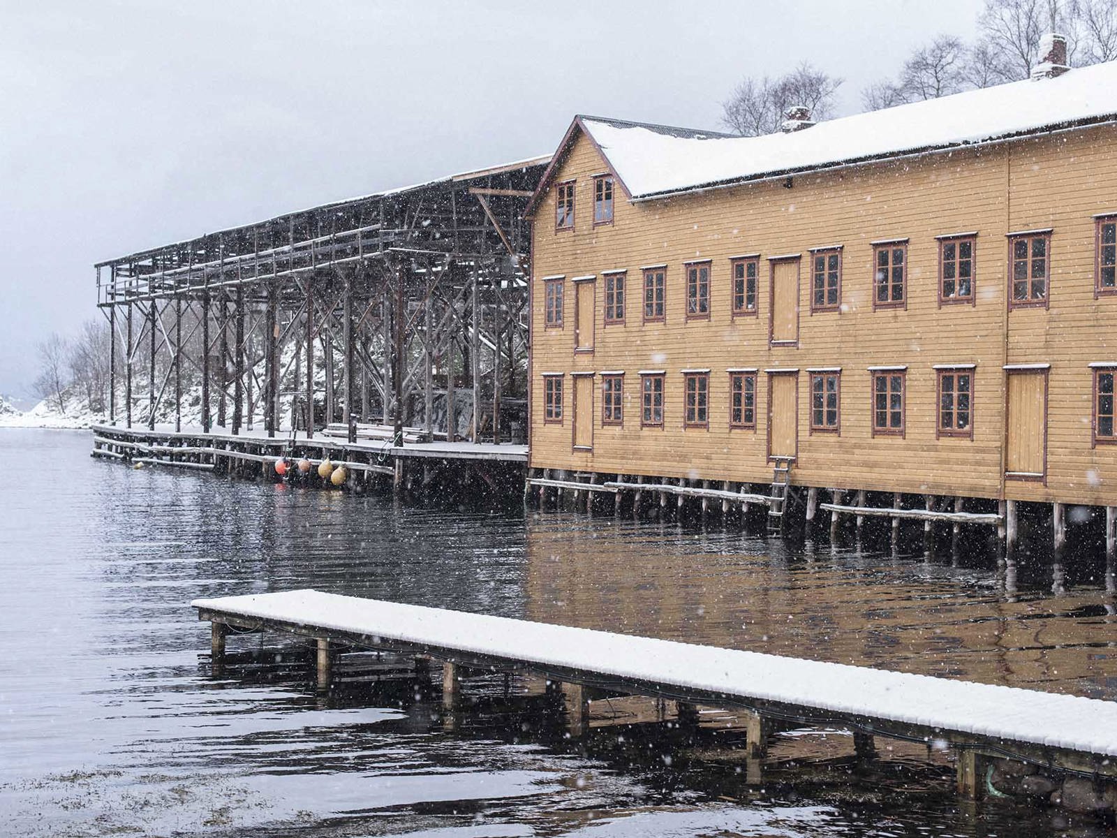 Gult hus inntil stort notheng bygg, alt på bryggekant. Brygge og vann i forgrunnen. Snø i lufta. Foto.