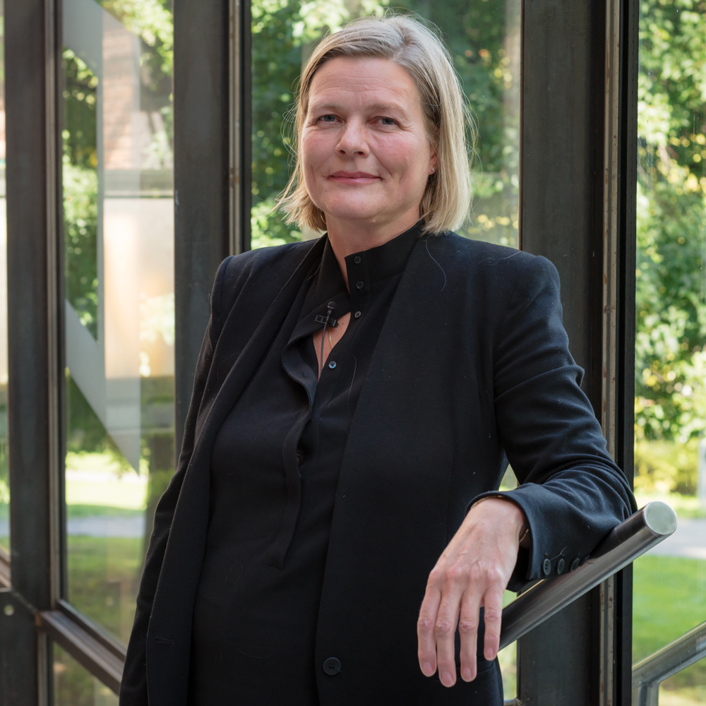 Marianne Skjulhaug lener seg mot et rekkverk, med vinduer som ser ut i det grønne i bakgrunnen. Foto.