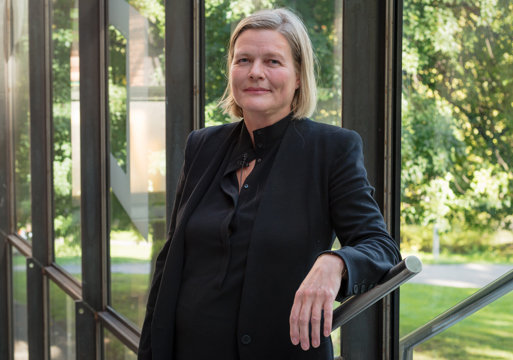 Marianne Skjulhaug lener seg mot et rekkverk, med vinduer som ser ut i det grønne i bakgrunnen. Foto.
