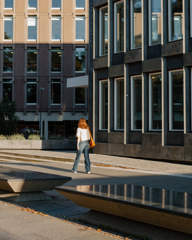 Foto av en kvinne som går i en gate med benker og gate i larvikitt, høye fasader rundt. 