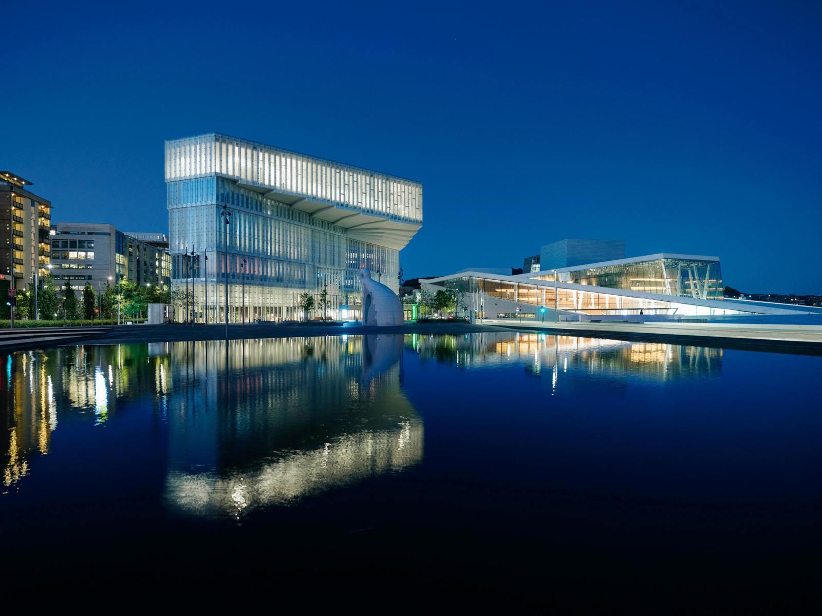 Foto av Deichmanske Bjørvika som speiles i vannet med Operaen på høyre side.