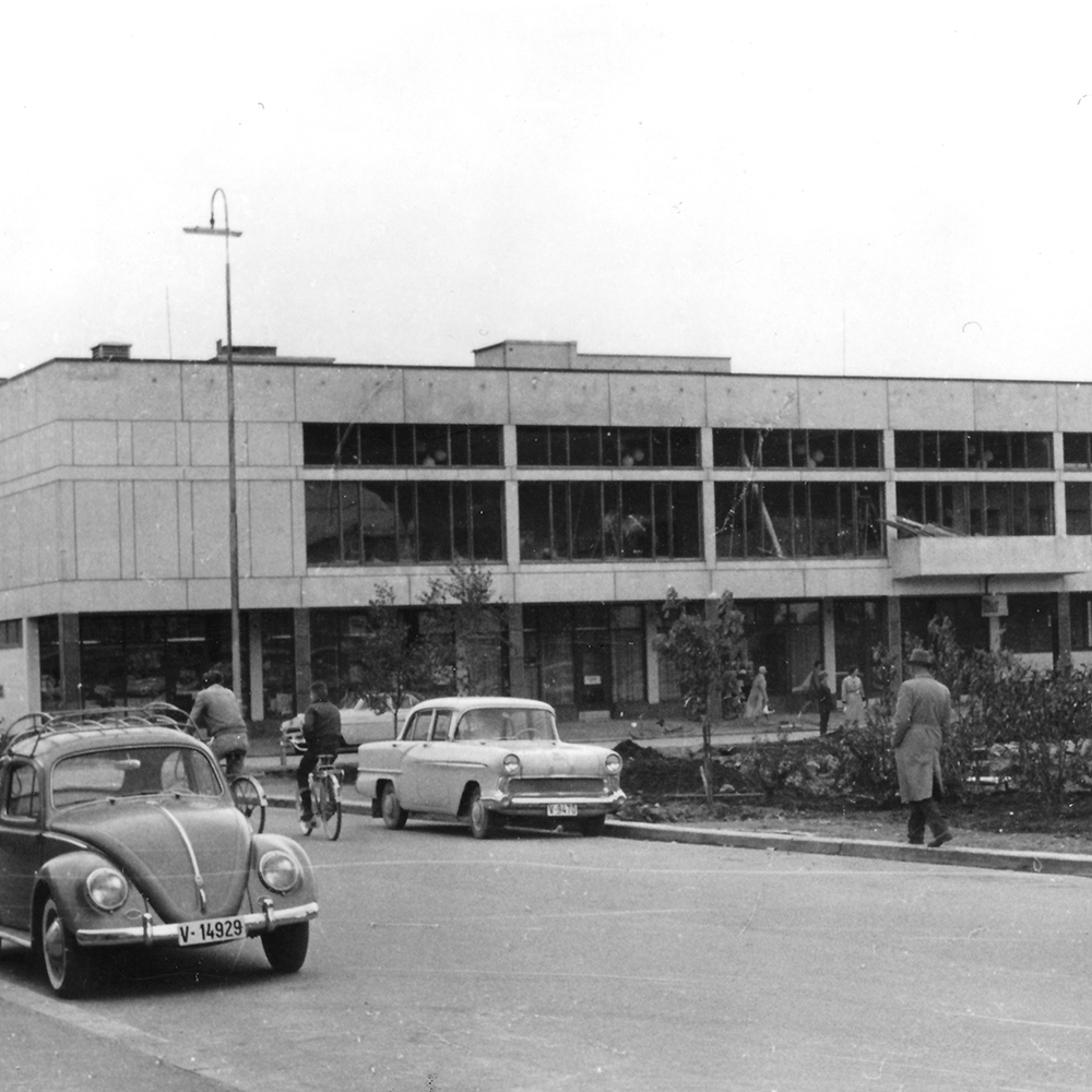 Modernistisk betongbygg og gamle biler som bobler kjører på veien, svart hvitt. Foto.