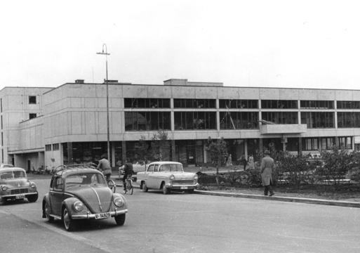 Modernistisk betongbygg og gamle biler som bobler kjører på veien, svart hvitt. Foto.