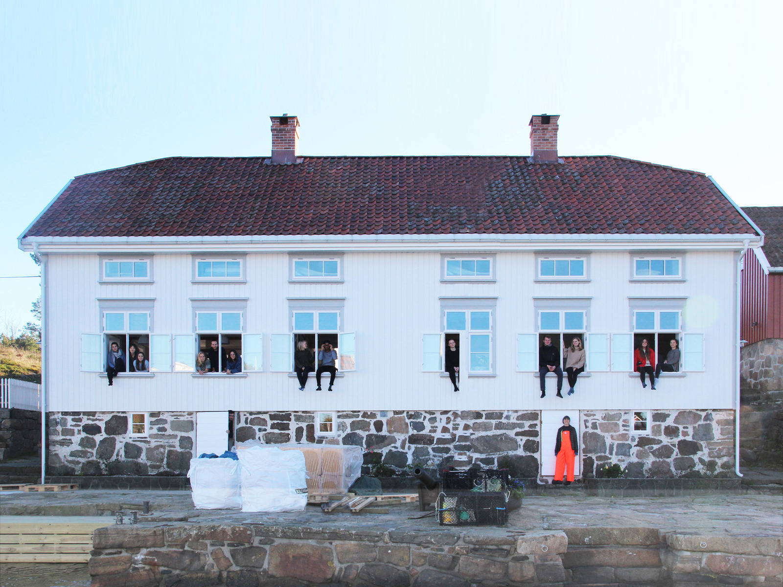 Hvit trehus med stein grunnmur på bryggekant, vann i forgrunnen. Flere mennesker sitter i vinduene i første etasje. Foto.