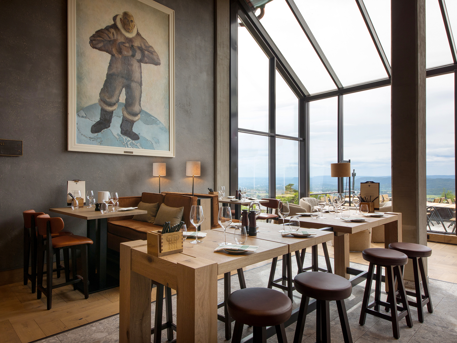 Ulike sittegrupper i restaurant/barområde med store vinduer og et portrett av Roald Amundsen på veggen. Foto.