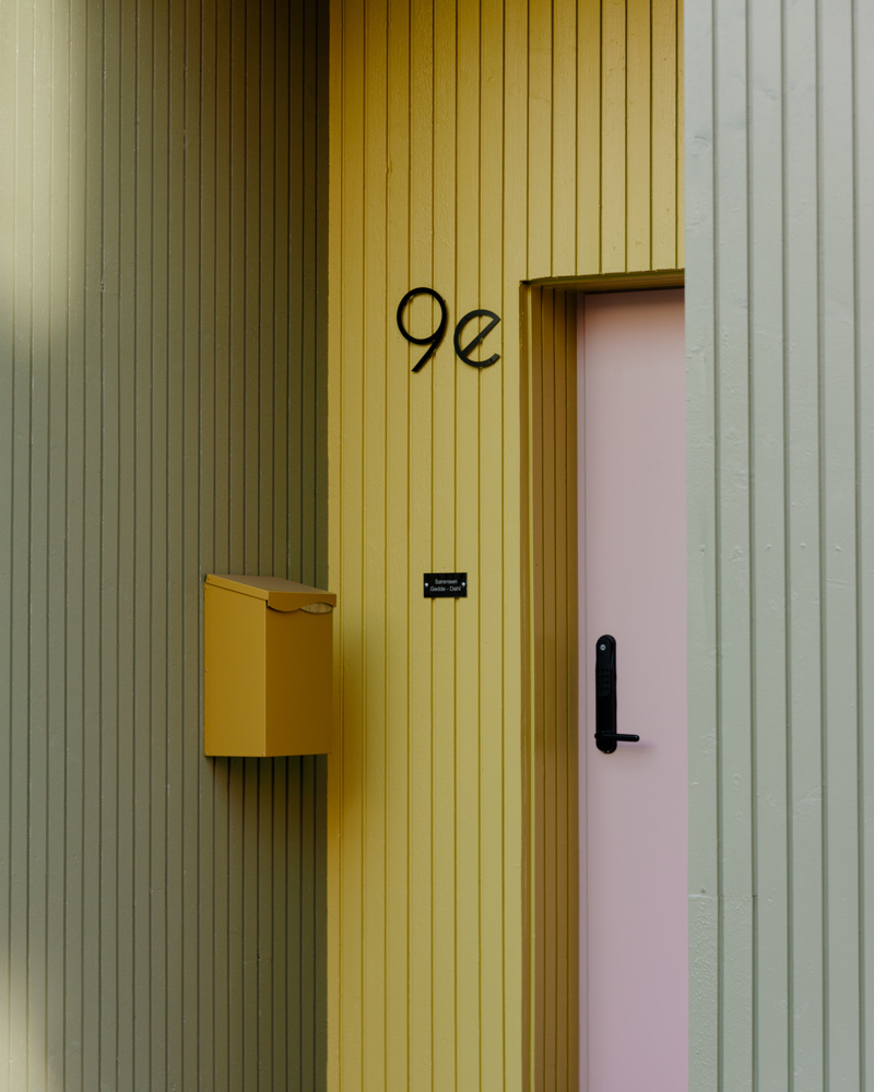 Rosa inngangsdør i en inntrukket nisje med gul postkasse og grønne vegger. Foto