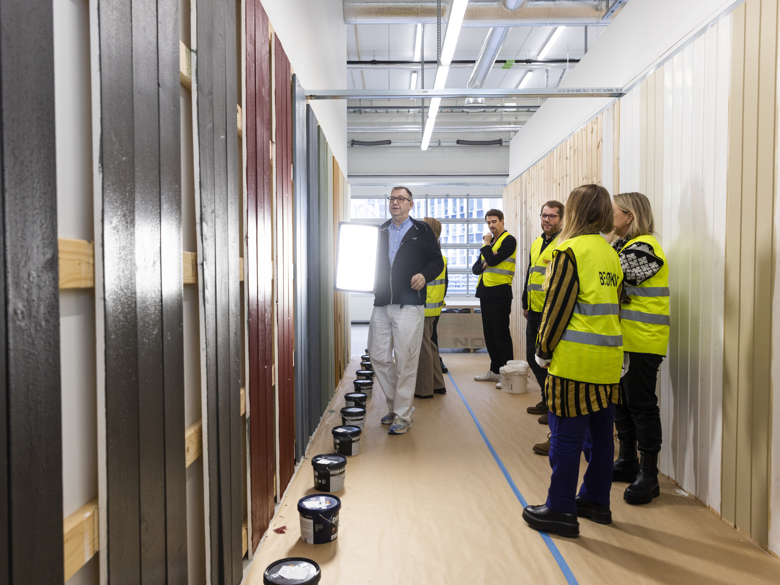 Vegg med malingsstester, lampe, mennesker i refleksvest. Foto.