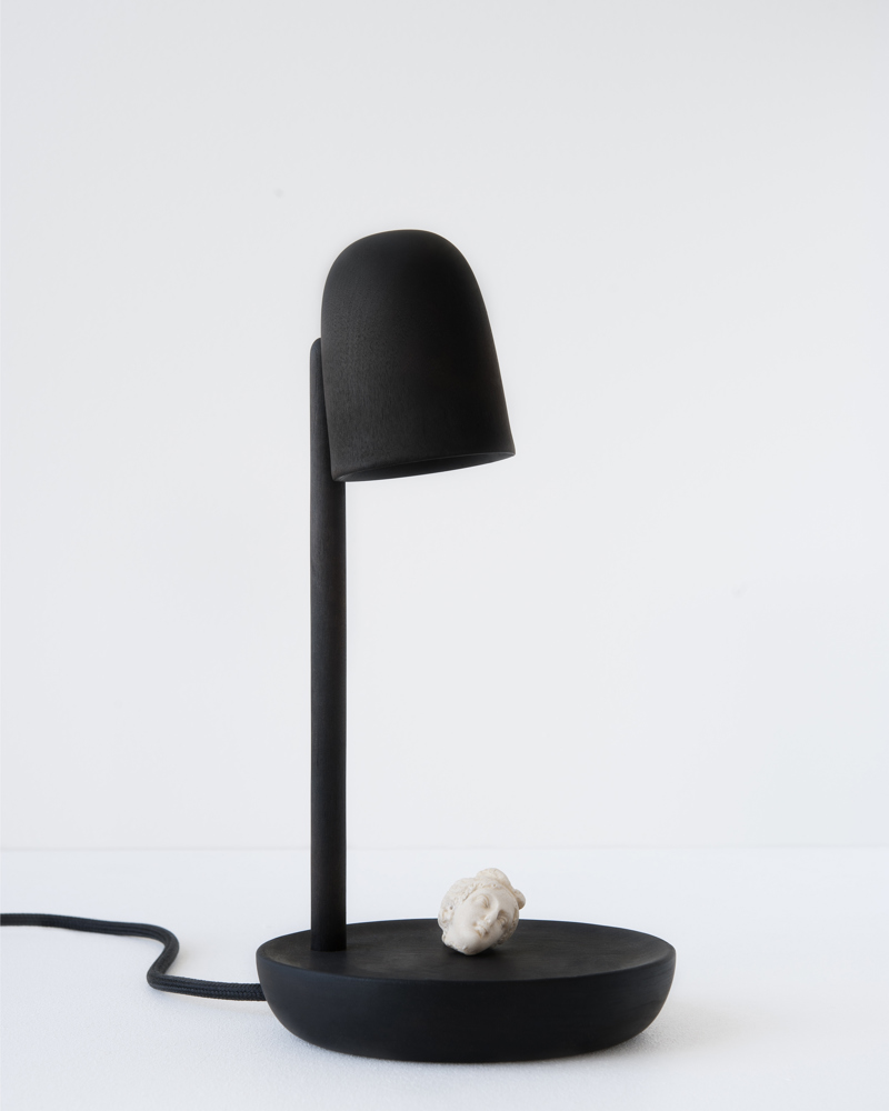 Produktbilde av en svart lampe med et rundt fat på bunn, der ligger det et hvitt lite gipshode. Foto.
