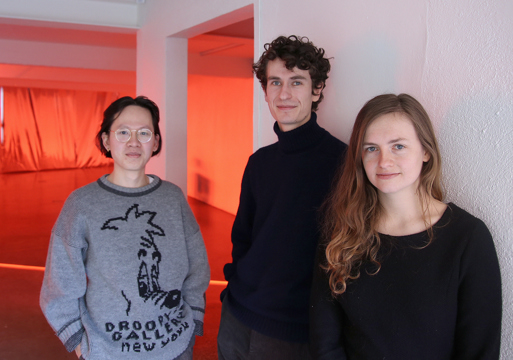 3 mennesker i et rom inntil en hvit vegg, rød lys fyller rommet i bakgrunnen. Foto.
