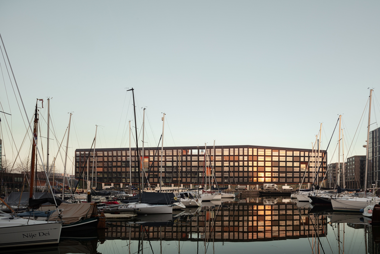 Foto av fasade på boligprosjekt som ligger ved havna, vann og båter fremfor, solnedgangslys.