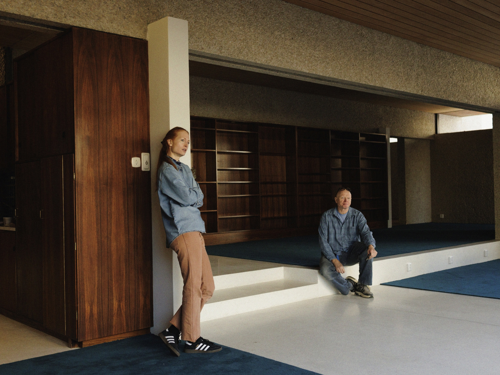 Interiør foto av en umøblert stue, en kvinne lener seg inntil veggen og en mann sitter i noen trappetrinn.