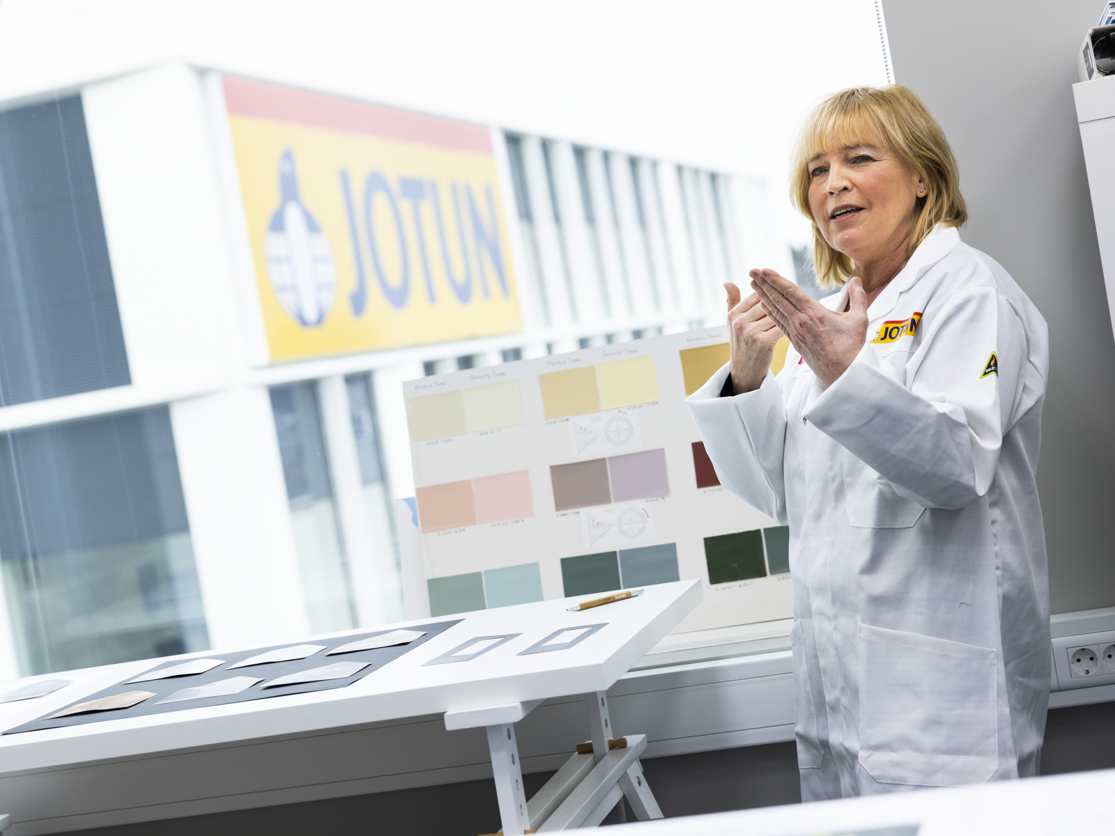 Kvinne i hvit frakk forklarer, fargeprøver på et bord. Foto.