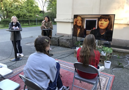 TV satt opp ute, 2 damer som står å snakker i hver sin mikrofon fremfor hvert sitt runde bord, klappestoler med publikum på et teppe. Foto.