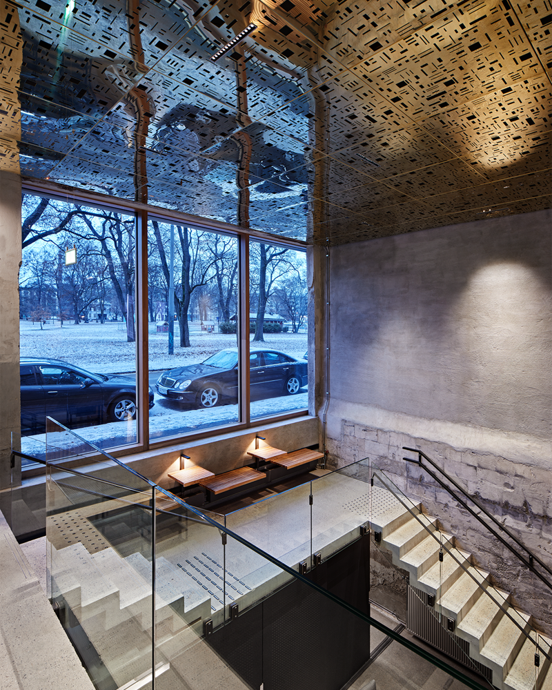 Interiør fotografi av bygg med flere sittegrupper, åpent til etasjen over og under, reflekterende gullplater i taket, betong trapp.