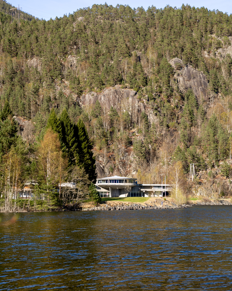Eksteriørfoto av kontorbygningen på Nesflaten tatt langt ifra for å vise hvordan anlegget ligger mellom mellom fjellet og Suldalsvatnet