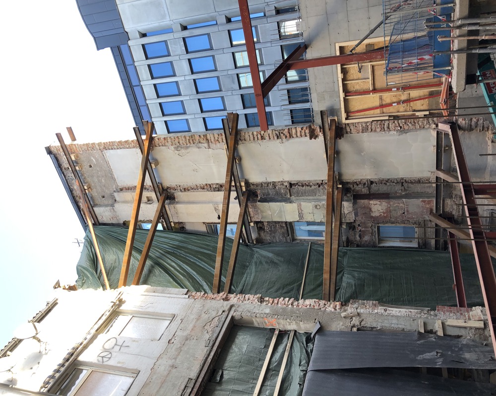 Eldre teglvegg står alene mellom bygg og holdes på plass av bjelker, byggeplass. Foto.