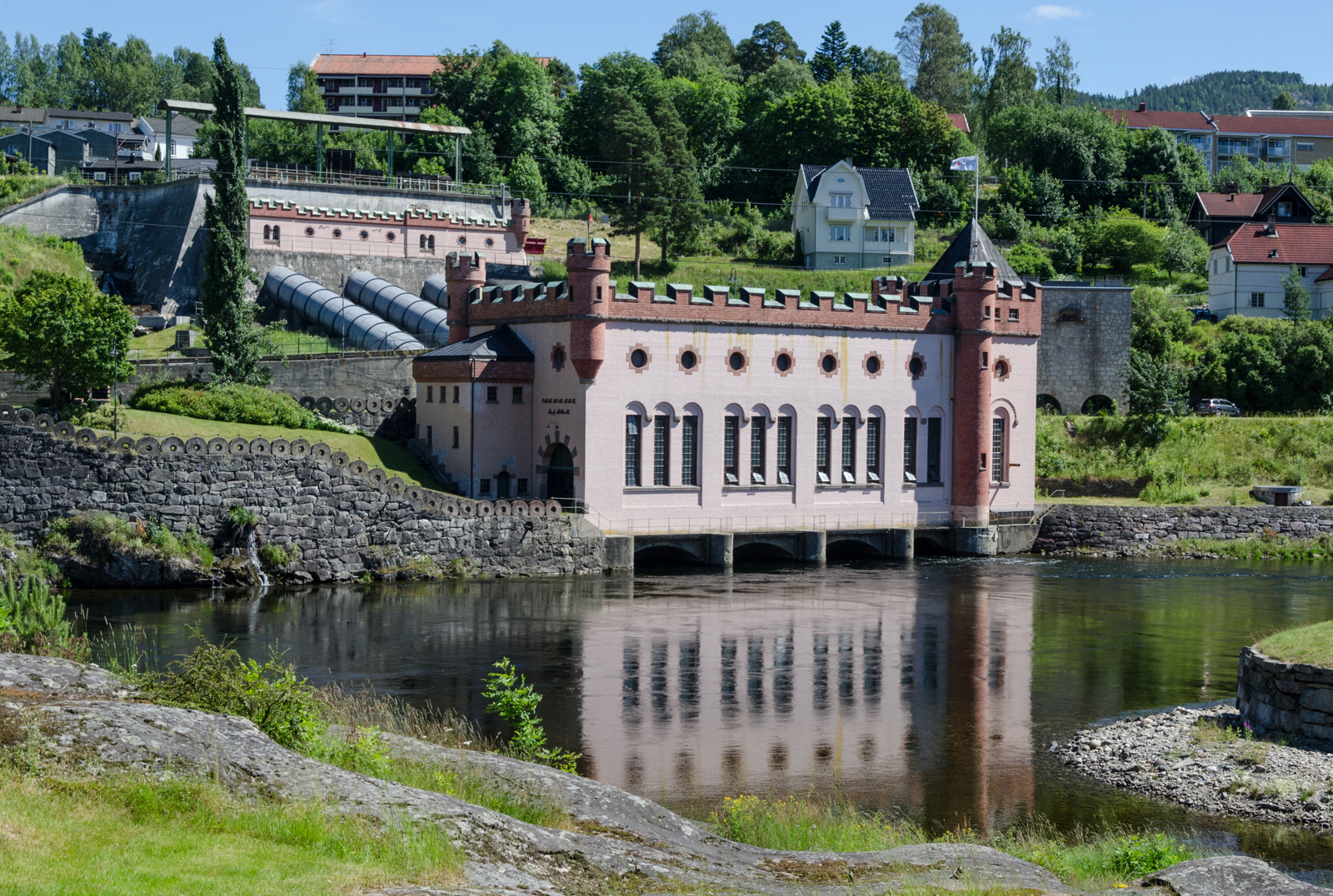 Foto av kraftverk i landskap, vann i forgrunnen, skog i bakgrunnen.