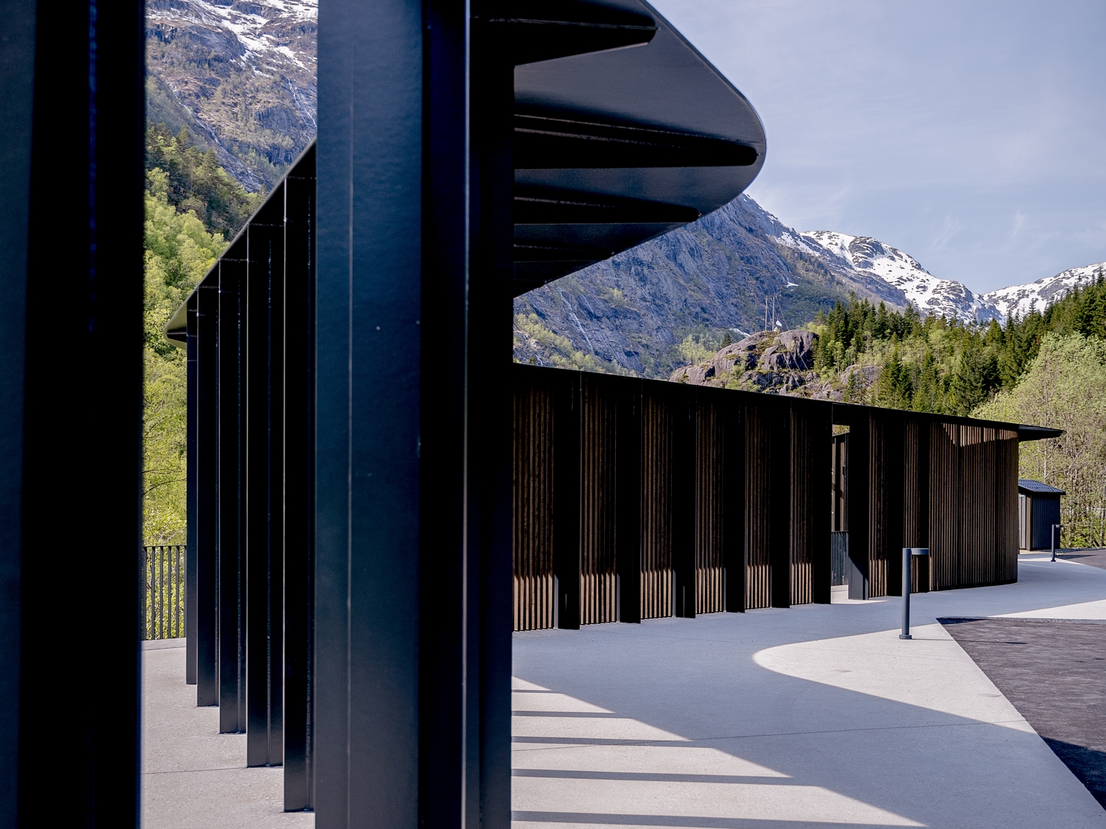 Mørk metallstruktur slynger seg over en parkeringsplass med fjell i bakgrunnen. Foto.