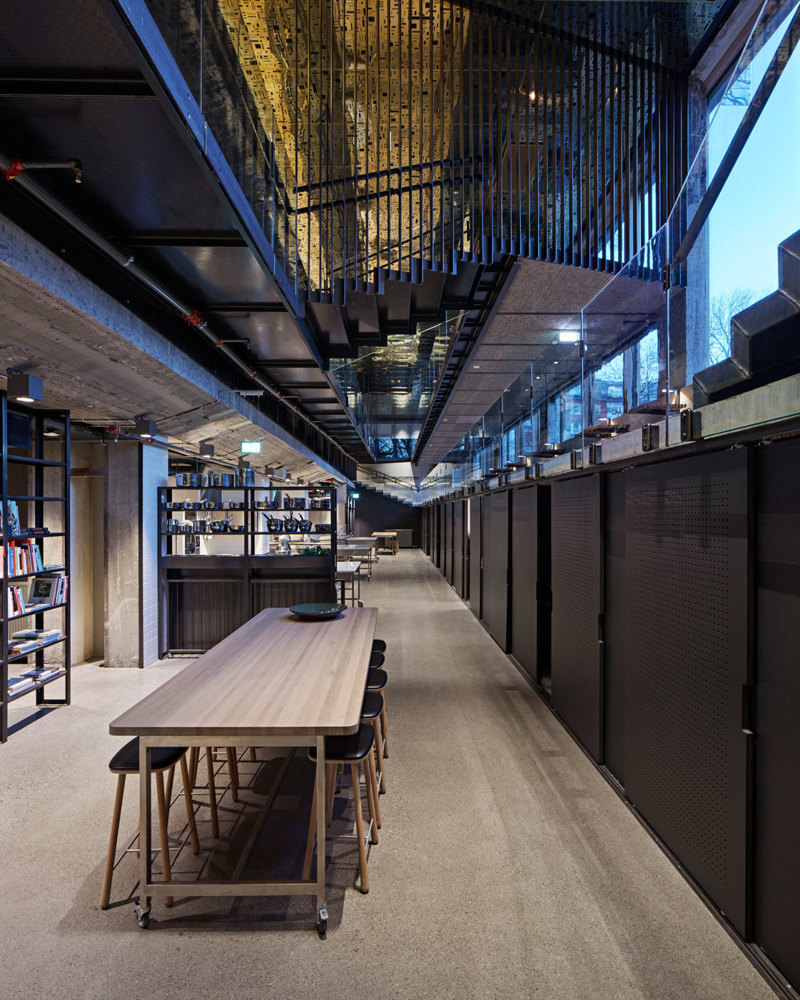 Interiør fotografi av bygg med flere sittegrupper, åpent ned til etasjen over, reflekterende gullplater i taket, ståltrapp med spiler.