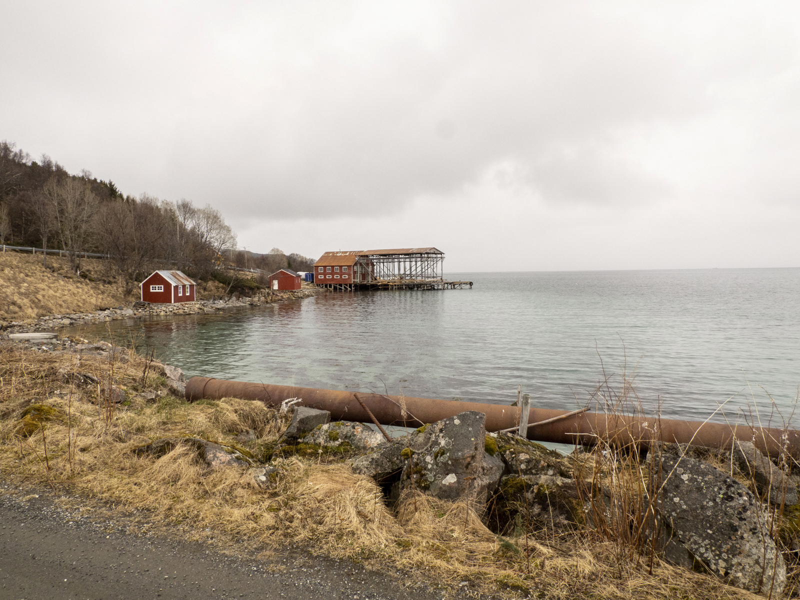 Røde hus langs kysten, notheng ut på brygge. veikant og rustent rør i forgrunnen. Foto.