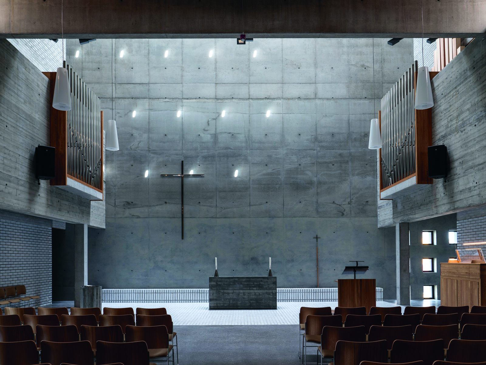 Kirkerom i betong, tre detaljer og stoler i tre. Overlys over alter. Foto.