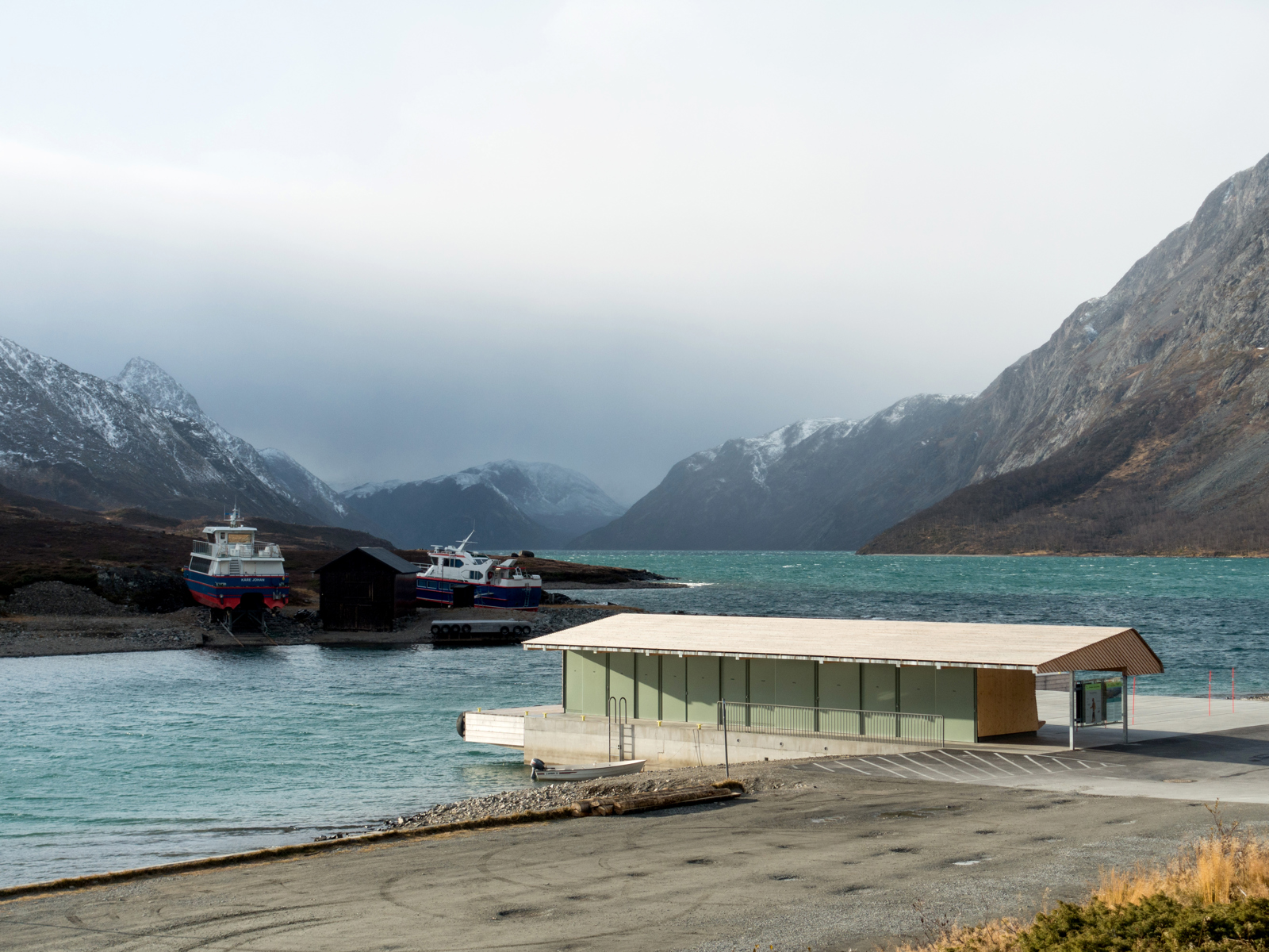 Bryggehus med vei fremfor, fjord og høge fjell i bakgrunnen. Foto.