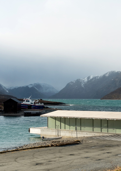 Bryggehus med vei fremfor, fjord og høge fjell i bakgrunnen. Foto.