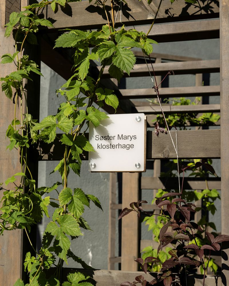 Foto av et skilt som henger på en hagestruktur med planter på seg. 