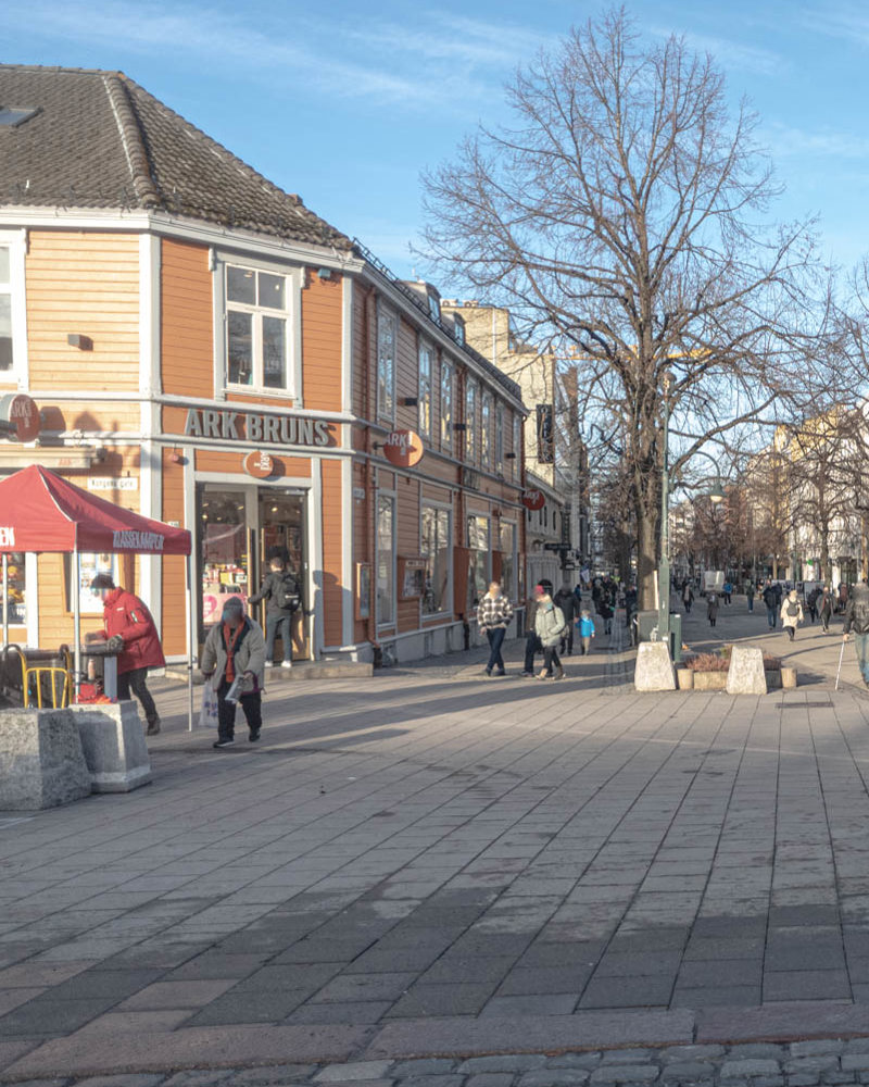 Torg plass i forgrunnen, trehus by i bak, folk som går rundt. Foto.