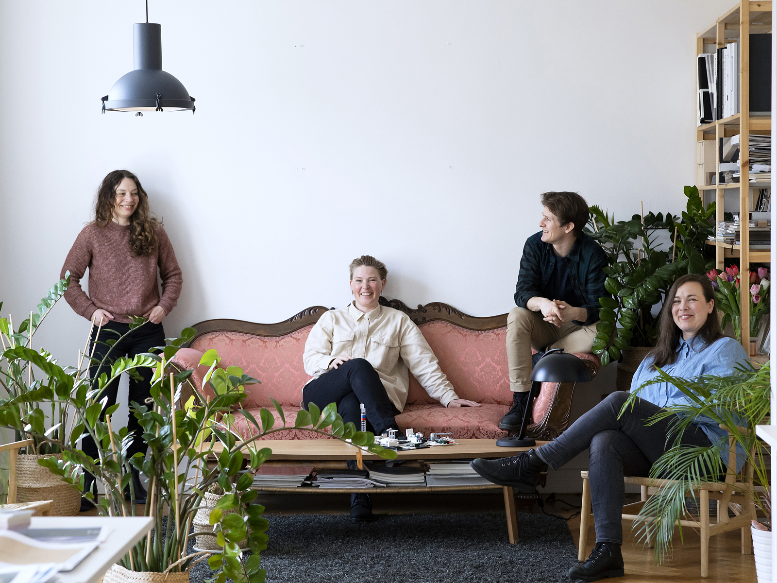 4 mennesker, en inntil veggen, en i en rosa sofa, en på kanten til sofarn og en i en stol. Foto.
