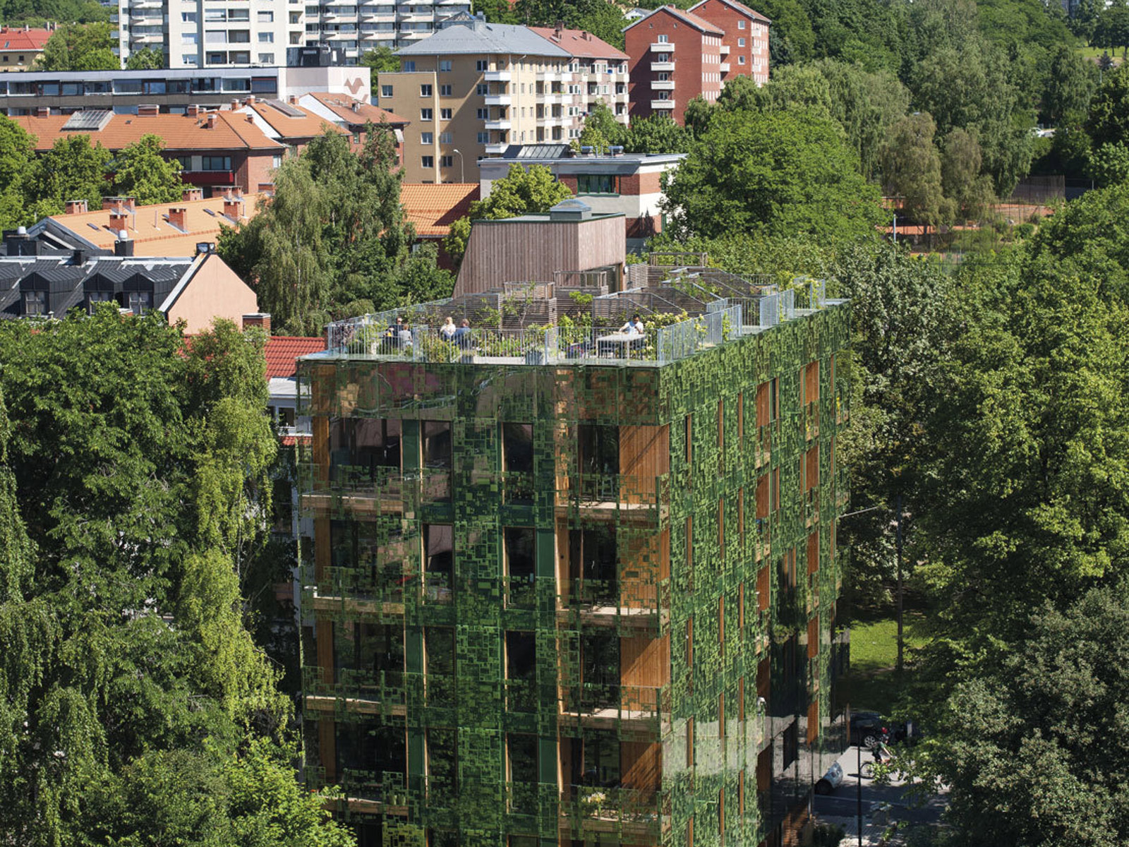 Grønn boligblokk i en kontekst med flere trær, by i bakgrunnen. Foto.
