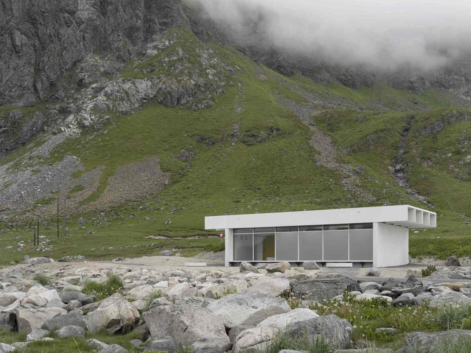 Foto av lite hvitt servicebygg midt i landskapet, fjell og skyer i bakgrunnen.