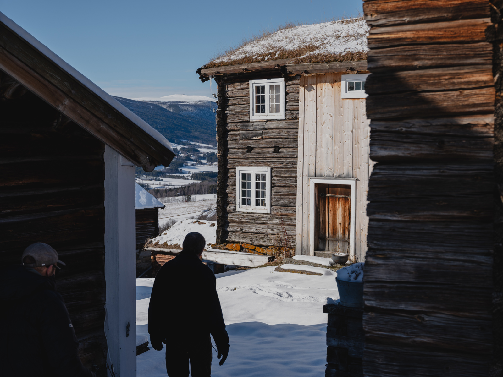 Foto av en mann som går mellom eldre hus på et gårdstun i vinterlandskap.