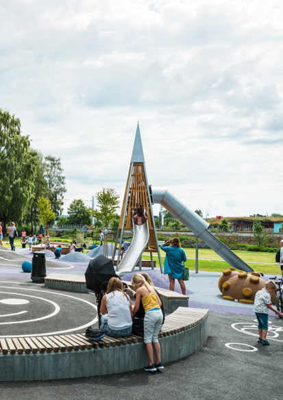 Foto av lekeplassen i Strandgateparken, benker og mange ulike lekeapparater, barn leker og voksne passer på.