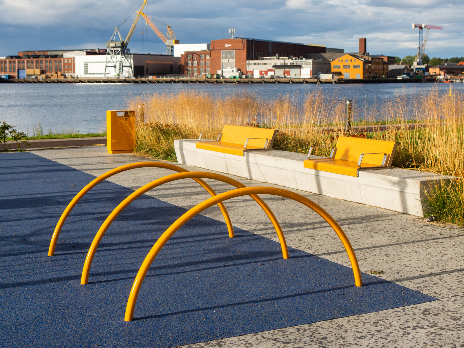 Sitteplasser og stor gule bøyler langs havnekanten, by og havn i bakgrunnen. Foto.