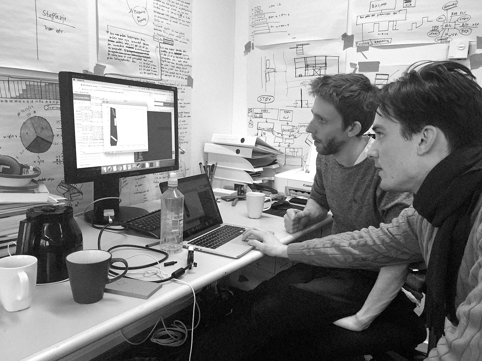 To menn sitter fremfor en pult og ser på en monitor, svart hvitt foto.