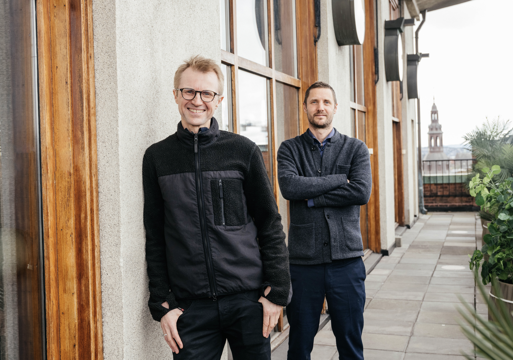 Bilde av to menn som står utenfor et hus, den ene lener seg inntil huset og den andre står med armene i kors. Foto.