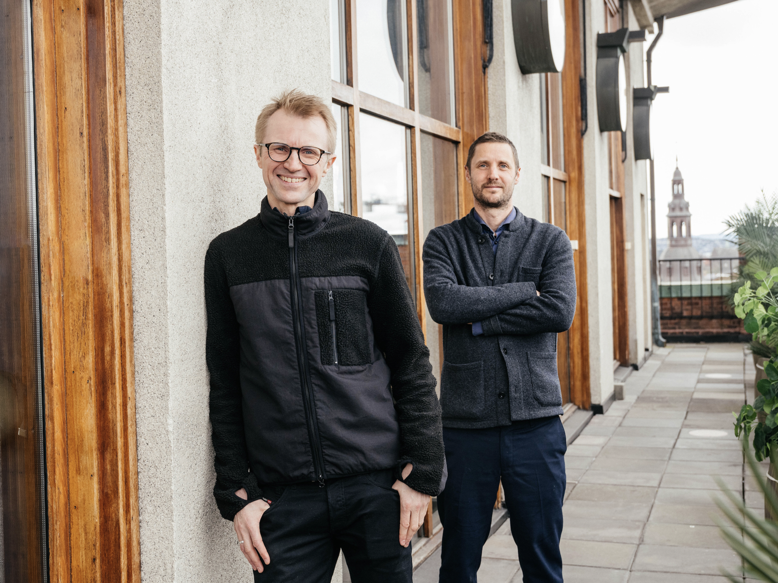Bilde av to menn som står utenfor et hus, den ene lener seg inntil huset og den andre står med armene i kors. Foto.