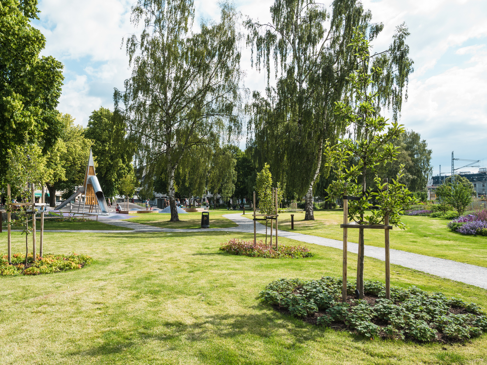 Foto av park med stor gressplen med plantede trær og grussti.
