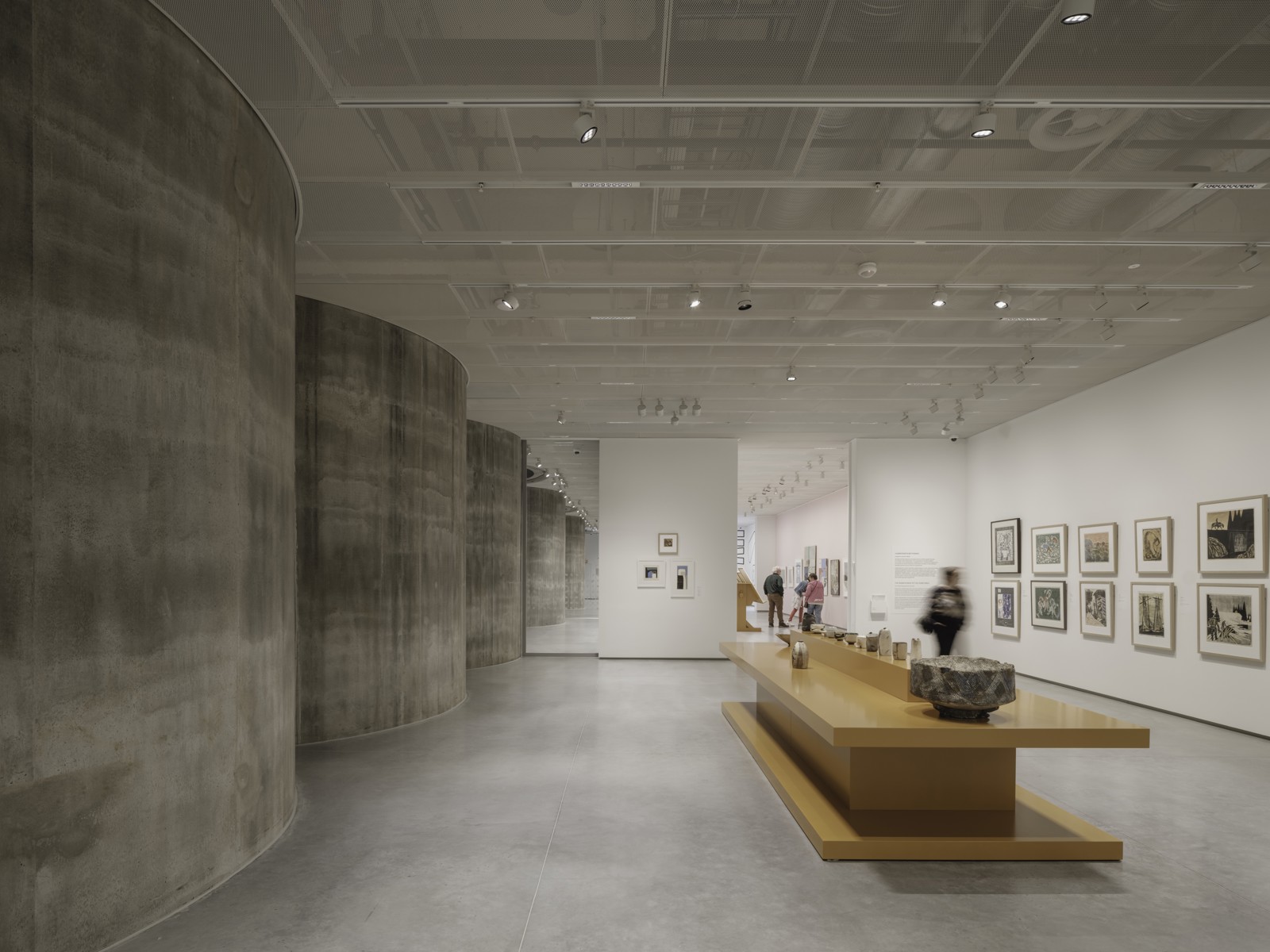 Interiør foto av utstillingsrom, oransje utstillingsbenk midt i rommet, røff betong vegg på venstre side og bilder stilt ut på en hvit blank vegg på høyre side.