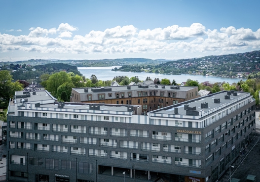 Oversiktsbilde av bolig kompleks, grå fasade, vann og åser i bakgrunnen. Foto. 