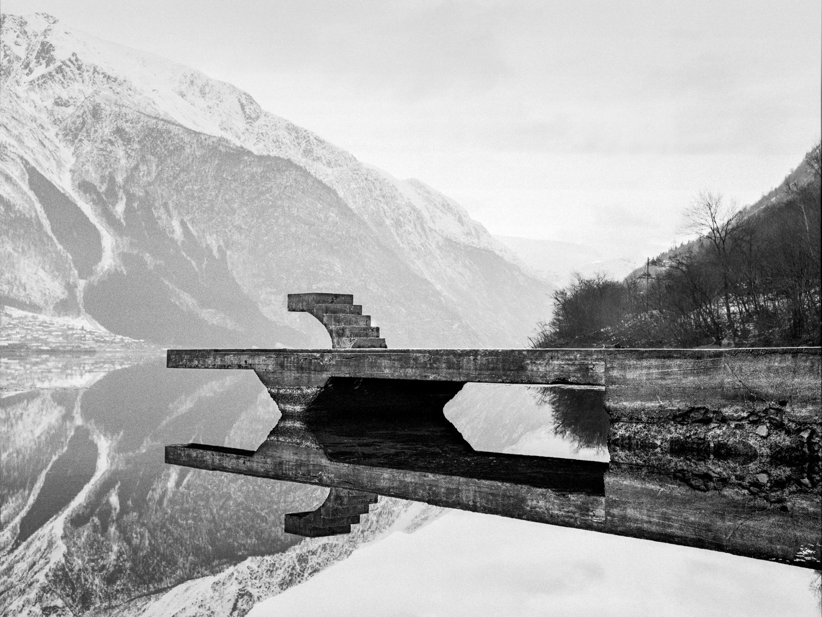 Svart hvitt fotografi av en brygge i betong med fjell ruvende i bakgrunn