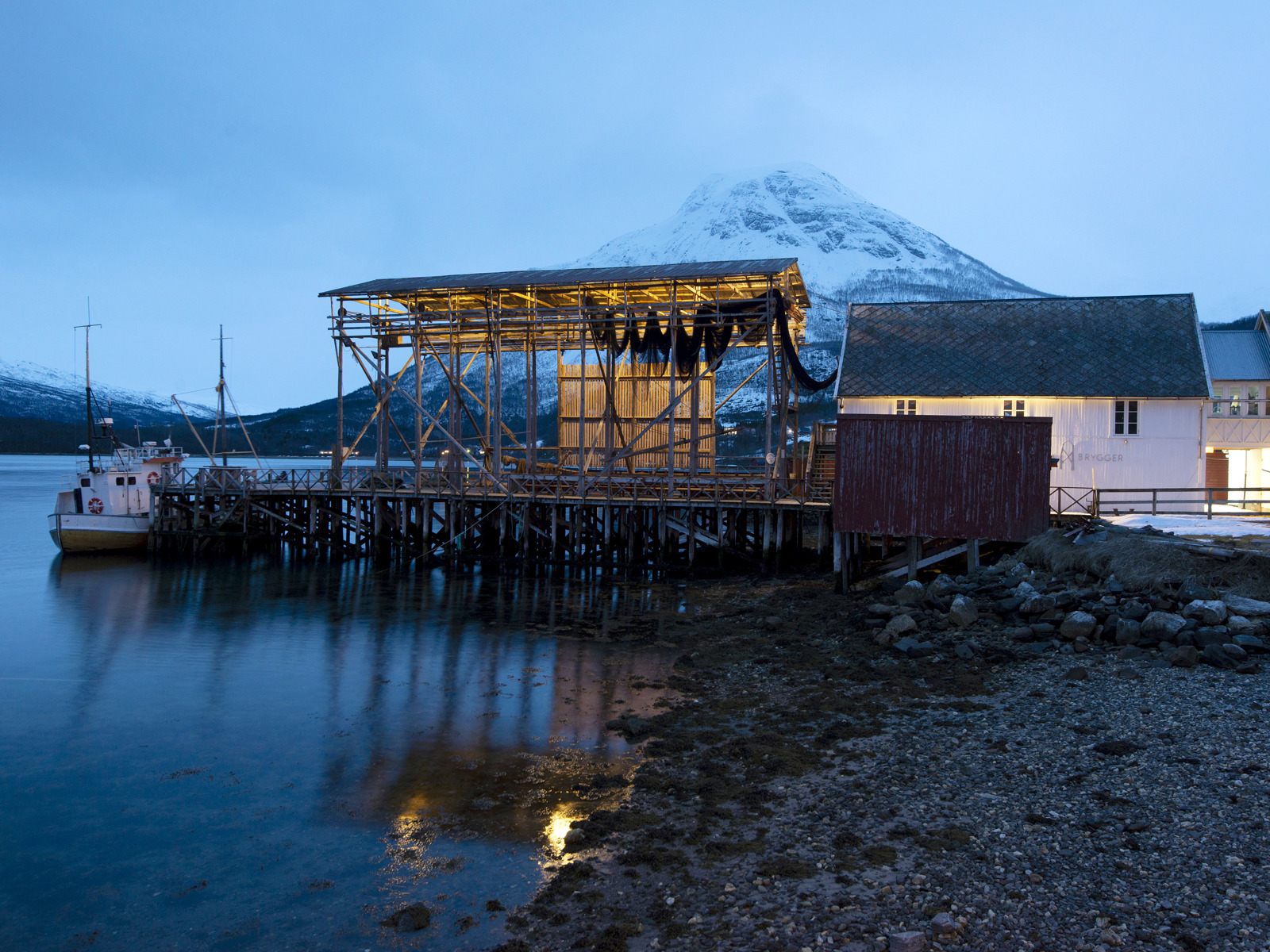 Kveldslys, blåtime, snøkledd fjell i bakgrunnen. Hvit hus, notheng på brygge, båt som ligger inntil bryggen, Vann i forgrunnen. Foto.