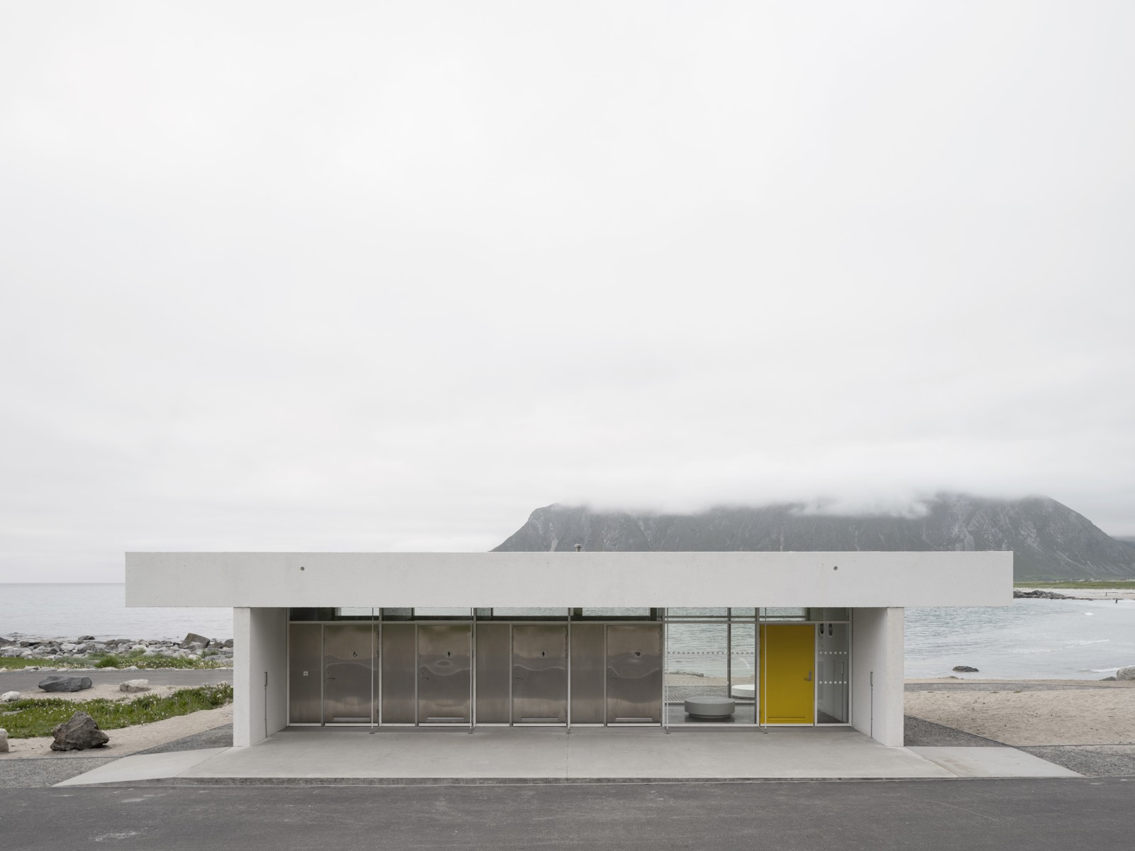 Foto av hvitt servicebygg midt i landskapet, hav, fjell og skyer i bakgrunnen.