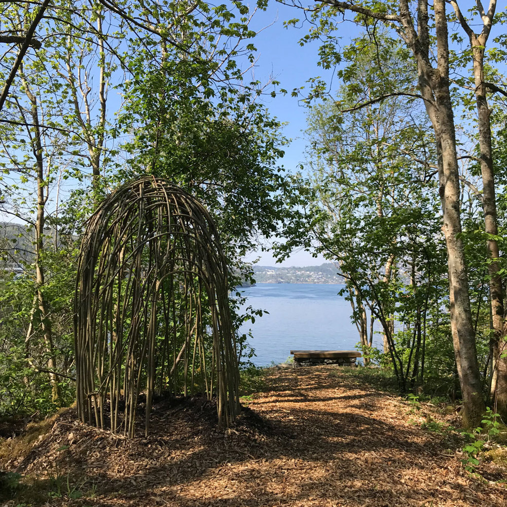 Bark på bakken, et rundt rom med dør inn laget av bøyde kvister. Utsikt mot havet. Foto.