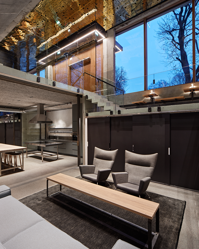 Interiør fotografi av bygg med flere sittegrupper, åpent til etasjen over, reflekterende gullplater i taket, betong trapp i bakgrunnen.