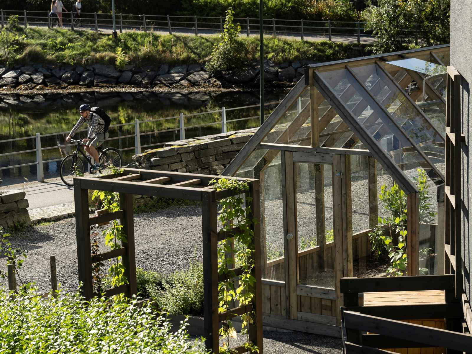 Foto av hage med drivhus og ulike strukturer, en gangvei med en mann som sykler, vann i bakgrunnen.