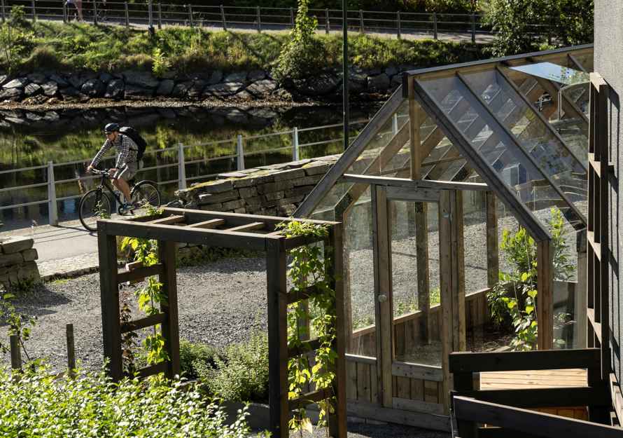 Foto av hage med drivhus og ulike strukturer, en gangvei med en mann som sykler, vann i bakgrunnen.