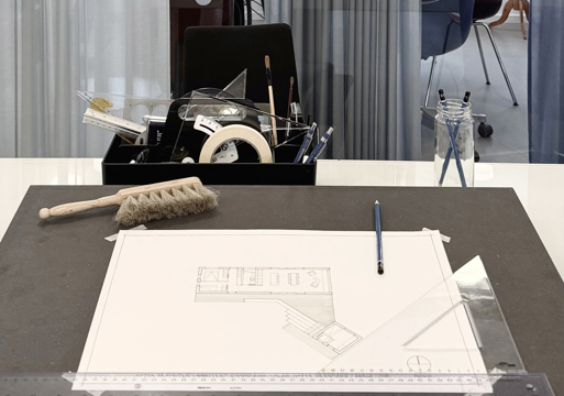 Foto av tegnebord med en arkitekt tegning og diverse tegneredskaper rundt.