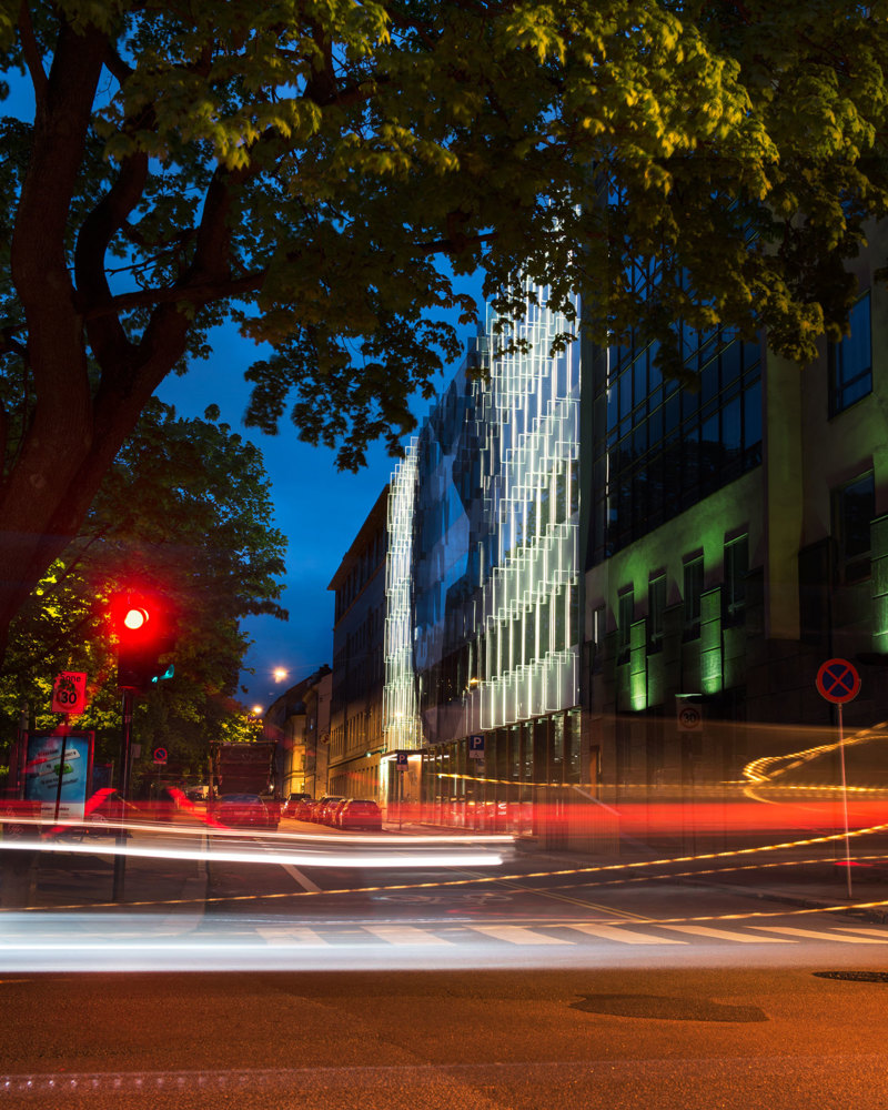 By fotografi i kveldsmørket, lysstriper fra biler i front, bygg med en opplysende fasade i bakgrunnen.